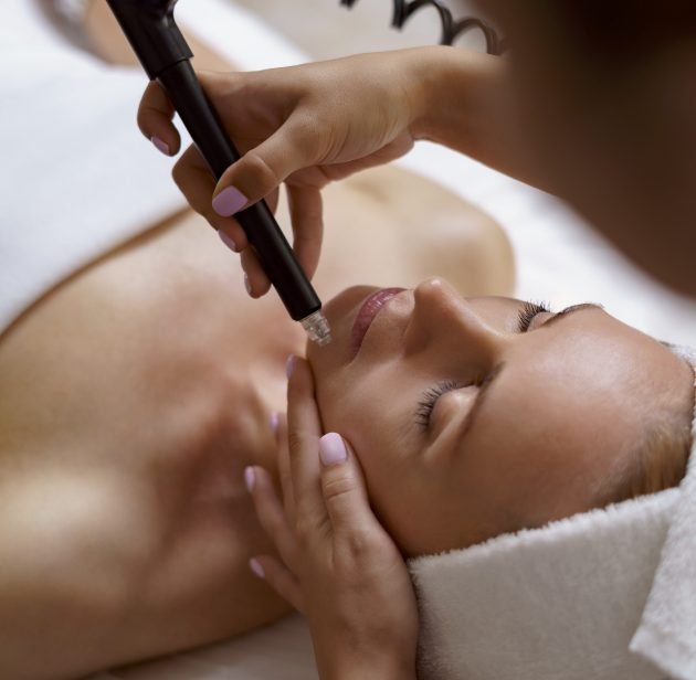 View of doctor cosmetologist doing anti aging procedure in cosmetology office. Satisfied woman in disposable hat lying on couch and relaxing. Working with Apparatus.